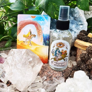 May include: A clear glass bottle with a black spray top and a white label that reads "Remember Your Divinity Aura Mist". The bottle is surrounded by white crystals and a tarot card with a woman riding a cheetah.