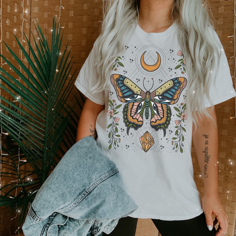 May include: White t-shirt with a colorful butterfly graphic, a crescent moon, and a crystal. The text "je ne regrette rien" is printed on the right side of the shirt.