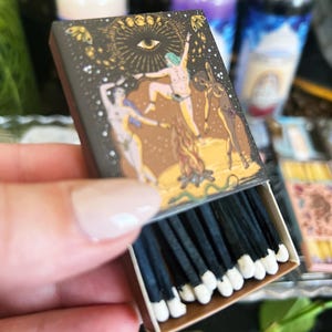 May include: A box of black matches with white tips. The box has a colorful illustration of a group of people dancing around a fire with a large eye in the sky. The text on the box reads "The Eye of the World".