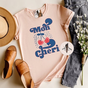 May include: A pink t-shirt with a blue and red graphic design that says "Moh Cheri" with two cherries.