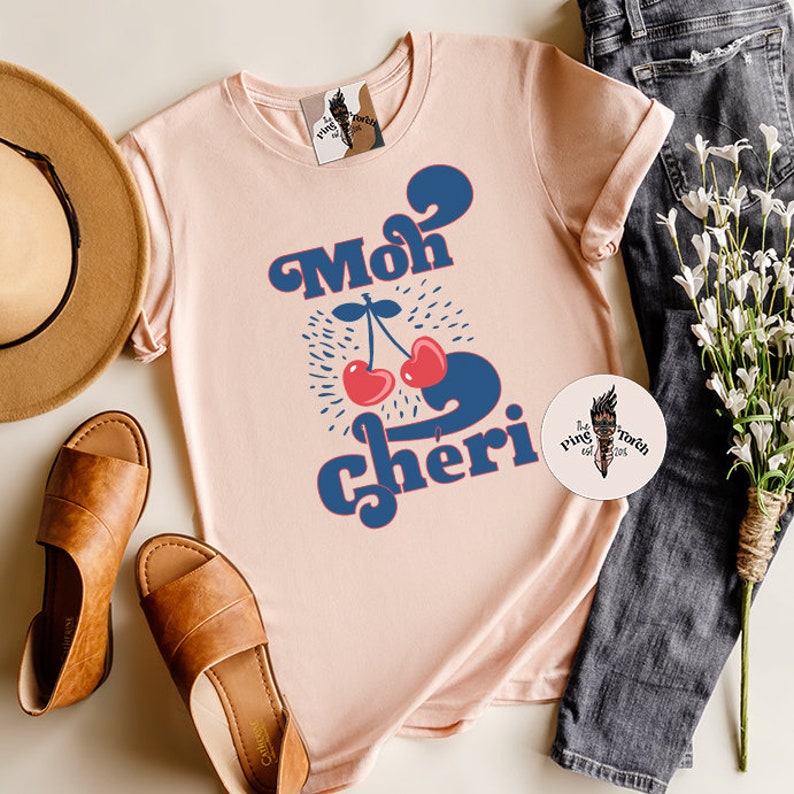 May include: A pink t-shirt with a blue and red graphic design that says "Moh Cheri" with two cherries.