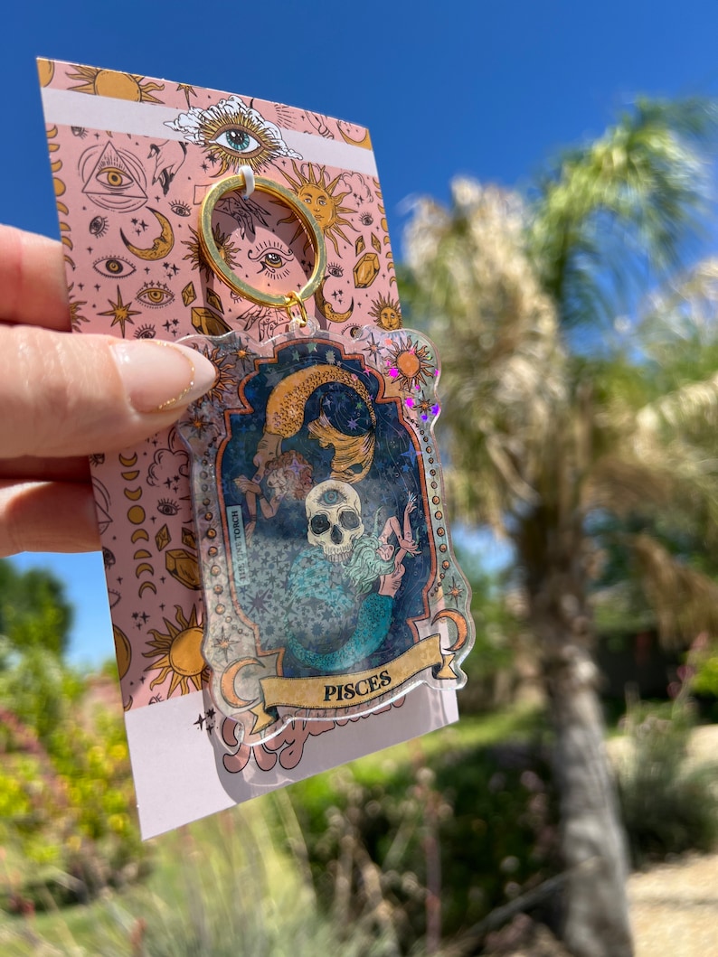 Puede incluir: Un llavero transparente con un marco dorado y una ilustraci&oacute;n colorida de una sirena y una calavera con la palabra "Pisces" en la parte inferior. El llavero est&aacute; sobre un fondo rosa con estrellas, lunas y ojos.