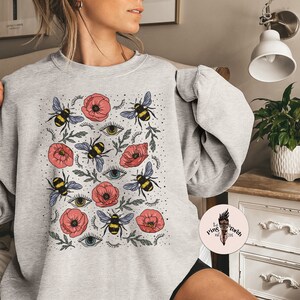 K&ouml;nnte beinhalten: Grauer Pullover mit einem Aufdruck von roten Mohnblumen, Bienen und stilisierten Augen. Der Aufdruck wiederholt sich auf dem gesamten Pullover.