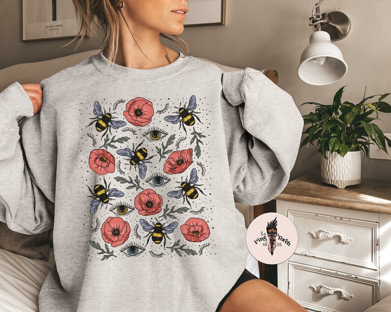 K&ouml;nnte beinhalten: Grauer Pullover mit einem Aufdruck von roten Mohnblumen, Bienen und stilisierten Augen. Der Aufdruck wiederholt sich auf dem gesamten Pullover.