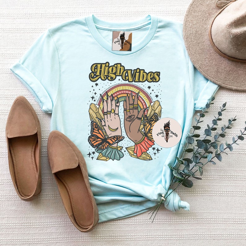 May include: Light blue t-shirt with the text "High Vibes" in a retro font. The design features two hands with crystals, butterflies, and a rainbow.
