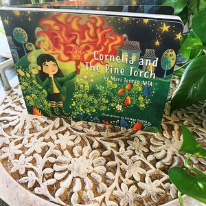 May include: A children's book titled "Cornelia and the Pine Torch" by Mari Toscas MFA. The cover features a girl holding a flaming torch in a green forest setting with a starry night sky.
