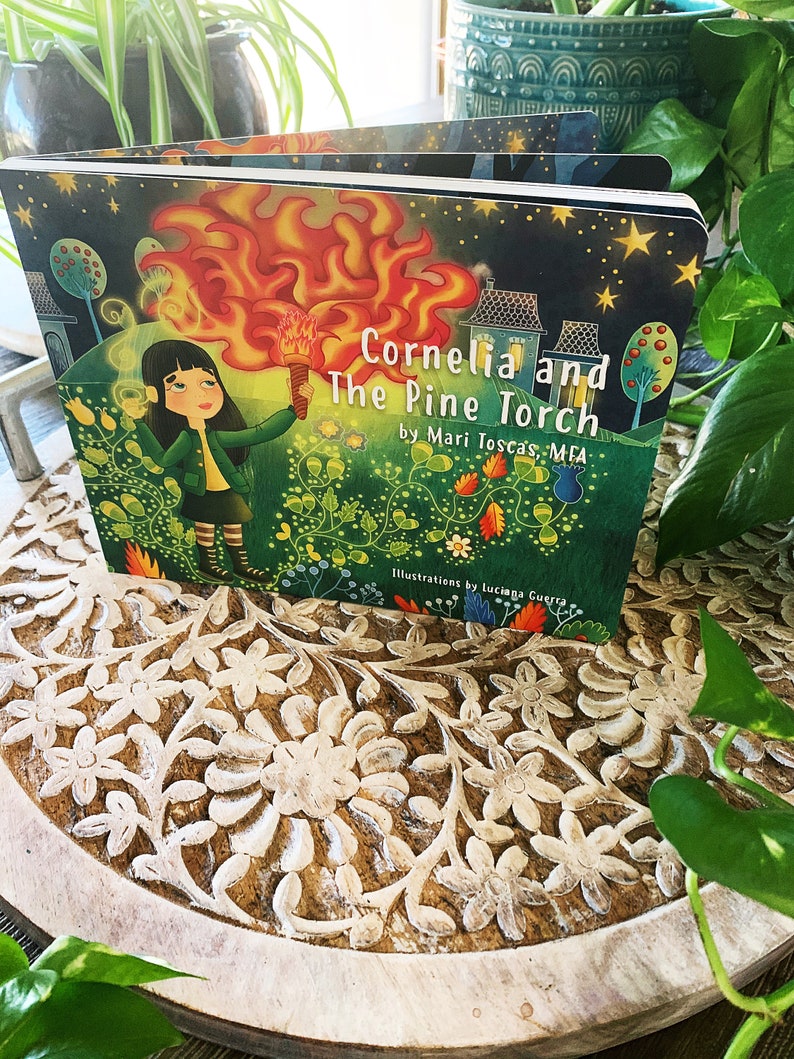 May include: A children's book titled "Cornelia and the Pine Torch" by Mari Toscas MFA. The cover features a girl holding a flaming torch in a green forest setting with a starry night sky.