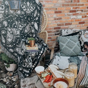 May include: A black and white tapestry with a moth and eye design draped over a wicker chair. A small potted plant sits on a table with a book titled "Mohawk's Dictionary" and a plate of food. Two glasses of iced coffee and a candle are on the table.