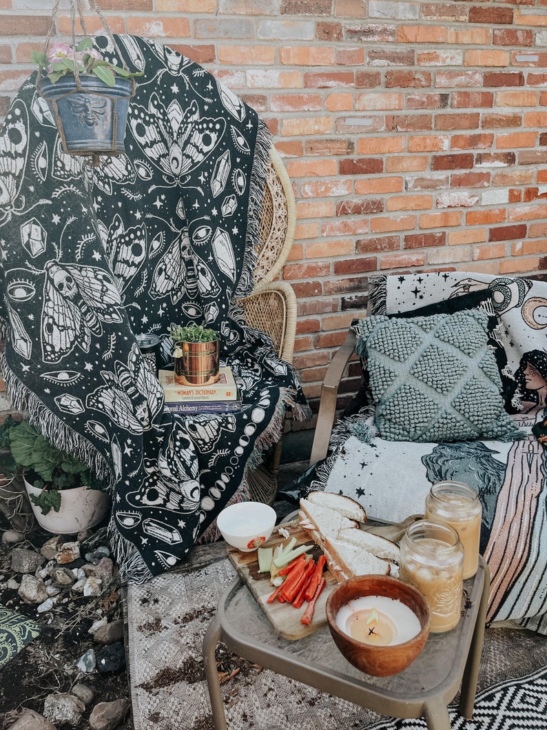 May include: A black and white tapestry with a moth and eye design draped over a wicker chair. A small potted plant sits on a table with a book titled "Mohawk's Dictionary" and a plate of food. Two glasses of iced coffee and a candle are on the table.