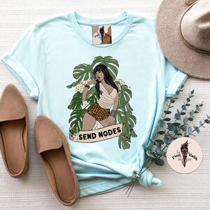 K&ouml;nnte beinhalten: Hellblaues T-Shirt mit einem Aufdruck einer Frau in einer Dschungelkulisse. Die Frau tr&auml;gt einen Bikini mit Leopardenmuster und ein wei&szlig;es Oberteil. Der Text "SEND NODES" ist unter dem Aufdruck gedruckt.