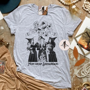 K&ouml;nnte beinhalten: Ein graues T-Shirt mit einer Schwarzwei&szlig;illustration von drei Hexen mit einem Kessel und dem Text "Mom Group Shenanigans".