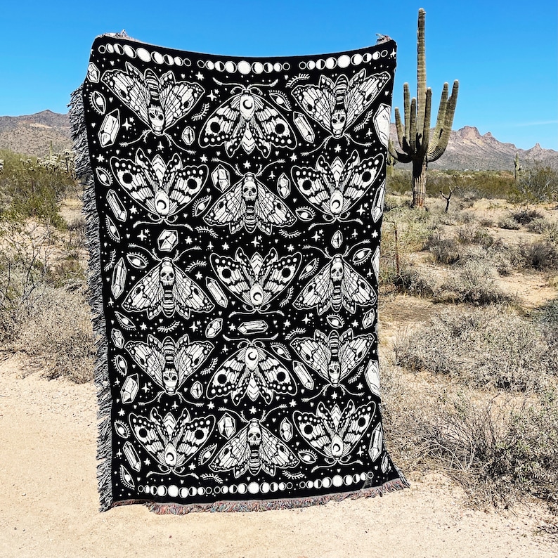 May include: Black and white woven throw blanket with a repeating pattern of moths, crystals, and eyes. The moths have skulls on their bodies and are surrounded by crescent moons.
