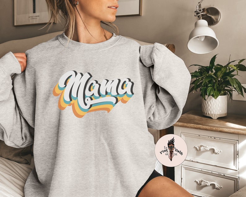 Pode incluir: Camisola cinzenta com um gr&aacute;fico de estilo retro que diz "Mama" em letras coloridas e sobrepostas.