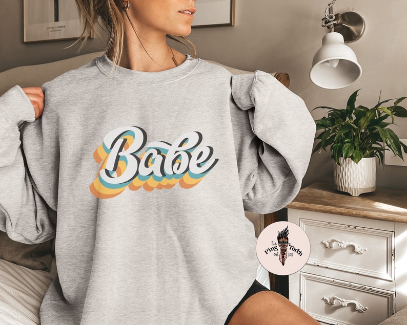 Może przedstawiać: Szara bluza z napisem "Babe" w retro tęczowym wzorze.