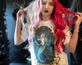 May include: White t-shirt with a vintage-style illustration of a mermaid with a Capricorn zodiac sign. The illustration is surrounded by stars and a crescent moon. The text "CAPRICORN" is printed below the illustration.