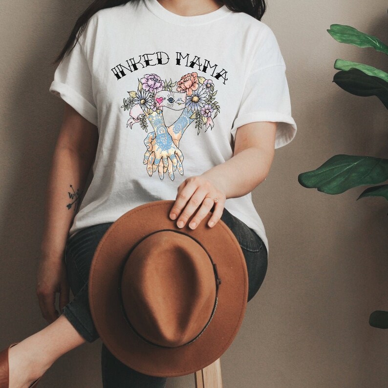 Pu&ograve; includere: T-shirt bianco con la scritta "INKED MAMA" sopra una grafica floreale e di mani colorata. Un cappello marrone &egrave; tenuto in grembo. Il design della maglietta presenta un cuore e fiori.