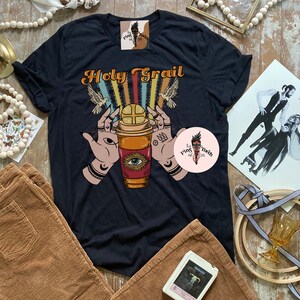 Op de afbeelding: Zwart T-shirt met een retro ontwerp met een koffiebeker met een oog, handen die de beker vasthouden en de tekst "Holy Grail".