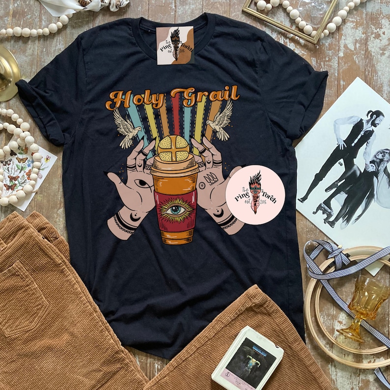 Op de afbeelding: Zwart T-shirt met een retro ontwerp met een koffiebeker met een oog, handen die de beker vasthouden en de tekst "Holy Grail".