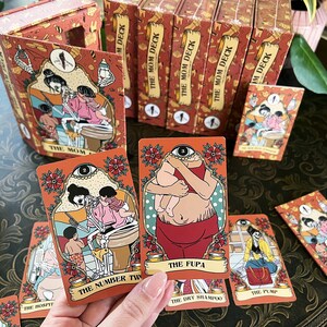 May include: A set of illustrated playing cards with a red, white, and black color scheme. The cards feature humorous illustrations of people and objects. The cards are titled "The Mom Deck".