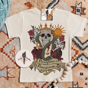 May include: A white t-shirt featuring a graphic print of a skull wearing a crown, a snake, and pomegranates. The text "Persephone's Pomegranates" is printed below the graphic.