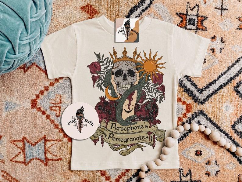 May include: A white t-shirt featuring a graphic print of a skull wearing a crown, a snake, and pomegranates. The text "Persephone's Pomegranates" is printed below the graphic.
