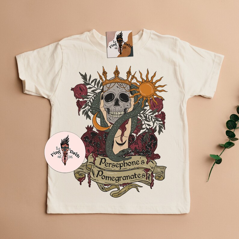 May include: A white t-shirt featuring a graphic design of a skull wearing a crown, surrounded by pomegranates and a snake. The text "Persephone's Pomegranates" is written on a banner below the skull.
