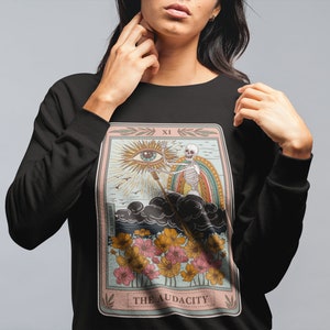 May include: Black sweatshirt with a tarot card design featuring a skeleton holding a staff with an eye at the top. The card is titled "The Audacity" and has a pink, blue, and yellow color scheme.