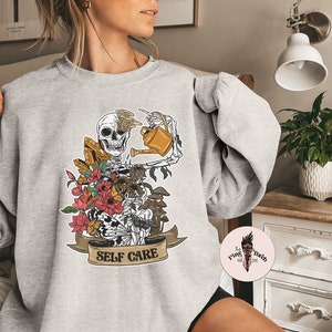 May include: A gray sweatshirt with a graphic of a skeleton watering flowers and mushrooms. The graphic has the text "SELF CARE" on a banner.