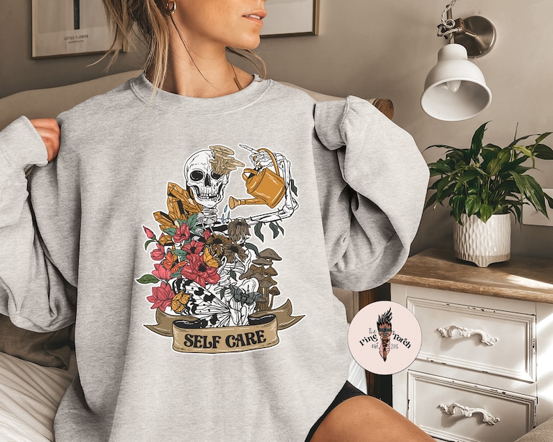 May include: A gray sweatshirt with a graphic of a skeleton watering flowers and mushrooms. The graphic has the text "SELF CARE" on a banner.