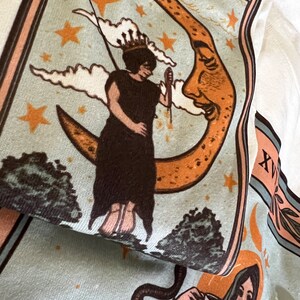 May include: A vintage-style illustration of a woman standing on a crescent moon with a face. The moon is orange and yellow with brown spots. The woman is wearing a long brown dress and a crown. The background is blue with white clouds and gold stars.