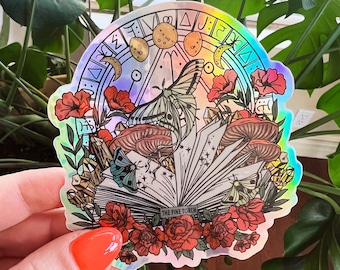 May include: A holographic sticker featuring a book with a butterfly, mushrooms, and flowers. The book is open and has the text "The Pine Torch" on the spine. The sticker has a rainbow iridescent background.