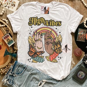 May include: White t-shirt featuring a graphic design of two hands with crystals and butterflies. The text "High Vibes" is printed in yellow on the shirt.