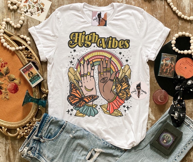 May include: White t-shirt featuring a graphic design of two hands with crystals and butterflies. The text "High Vibes" is printed in yellow on the shirt.