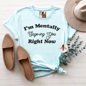 May include: A light blue t-shirt with the text "I'm Mentally Sage-ing You Right Now" printed in black. The shirt is laid out on a white surface with brown shoes and a straw hat.