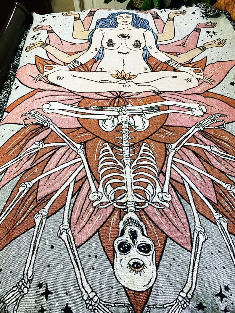 May include: A tapestry featuring a multi-armed goddess figure sitting in a lotus position atop a skeletal figure with a skull. The goddess has a third eye and is surrounded by stars. The tapestry is woven with a variety of colors, including pink, brown, and gray.
