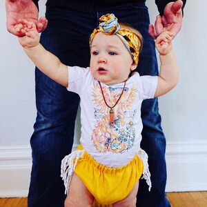 May include: A baby wearing a white onesie with a phoenix graphic and the text "Fight Like a Girl". The baby is also wearing a yellow skirt with fringe, a brown necklace with a tassel, and a blue and yellow headband.