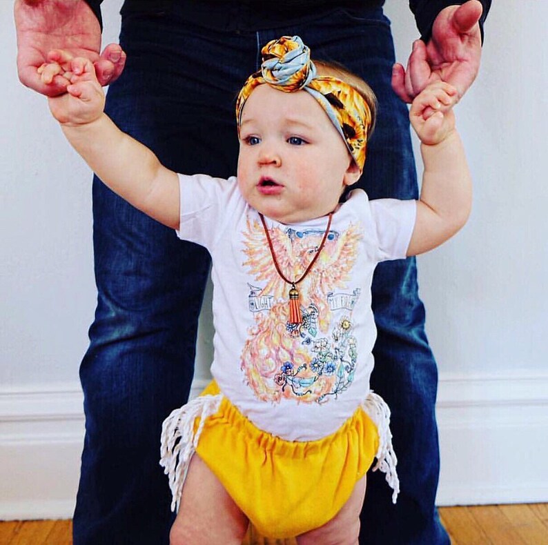 May include: A baby wearing a white onesie with a phoenix graphic and the text "Fight Like a Girl". The baby is also wearing a yellow skirt with fringe, a brown necklace with a tassel, and a blue and yellow headband.