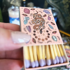 Könnte beinhalten: Eine rosa Streichholzschachtel mit einer bunten Illustration einer Schlange, Kristallen und Augen. Die Streichhölzer haben violette Spitzen.
