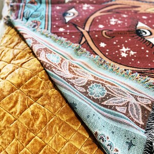 May include: A turquoise and brown woven throw blanket with a red background featuring a large eye design and white stars. The blanket is folded and draped over a brown quilted surface.