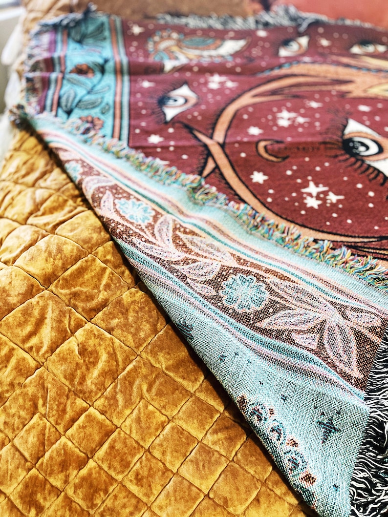 May include: A turquoise and brown woven throw blanket with a red background featuring a large eye design and white stars. The blanket is folded and draped over a brown quilted surface.