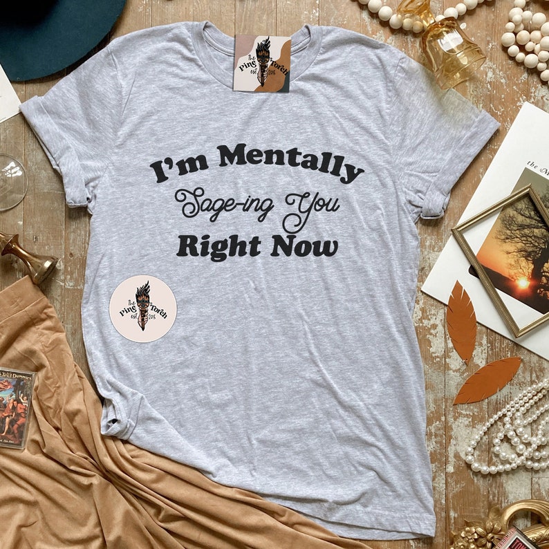 May include: A gray t-shirt with the text "I'm Mentally Sage-ing You Right Now" printed in black. The shirt is laid flat on a wooden surface with other decorative items.