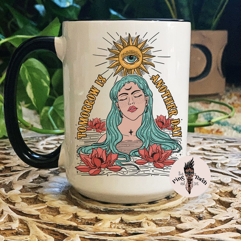May include: White ceramic mug with a black handle. The mug features a colorful illustration of a woman with long blue hair, a sun with an eye, and the text "Tomorrow is another day".