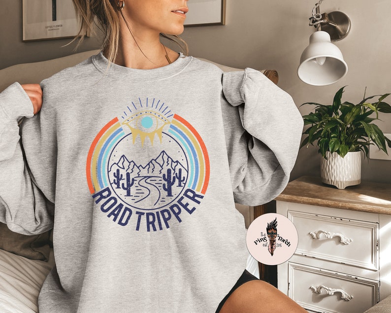 Peut inclure: Sweat-shirt gris avec un graphique r&eacute;tro repr&eacute;sentant une cha&icirc;ne de montagnes, une route sinueuse et un &oelig;il stylis&eacute; avec un arc-en-ciel. Le texte "ROAD TRIPPER" est imprim&eacute; sous le graphique.