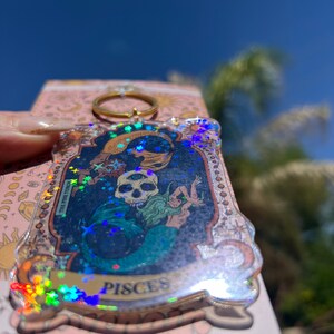 Puede incluir: Un llavero transparente e iridiscente con un anillo dorado. El llavero presenta una imagen hologr&aacute;fica de una sirena con una calavera y el texto "Pisces" y "The Pine Touch".