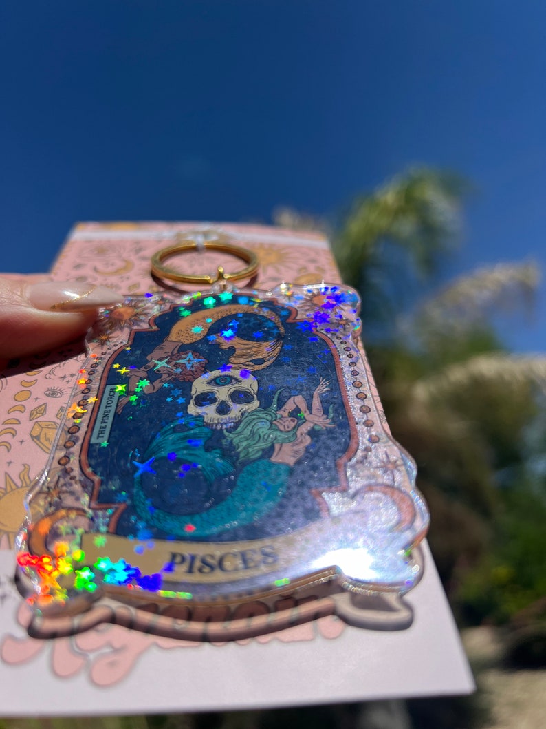 Puede incluir: Un llavero transparente e iridiscente con un anillo dorado. El llavero presenta una imagen hologr&aacute;fica de una sirena con una calavera y el texto "Pisces" y "The Pine Touch".
