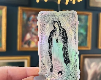 May include: A holographic sticker featuring a black and white line drawing of the Virgin Mary with a crown, standing on a crescent moon. The image is surrounded by a floral border and the text "The Fine Torch" is visible in the lower left corner.