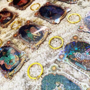 Puede incluir: Un conjunto de doce llaveros del zod&iacute;aco con purpurina hologr&aacute;fica y detalles dorados. Cada llavero presenta un signo del zod&iacute;aco diferente con su nombre correspondiente.