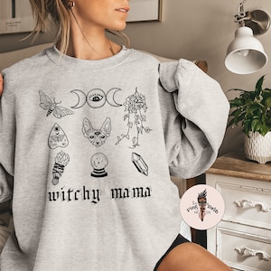 May include: Gray sweatshirt with a black and white graphic print of various witchy symbols, including a crescent moon, a cat, a crystal, and a moth. The text "witchy mama" is printed below the symbols.
