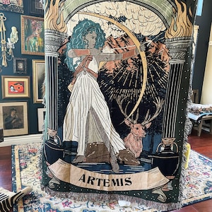 May include: A tapestry featuring a green background with a depiction of Artemis, the Greek goddess of the hunt, holding a bow and arrow. She is standing in front of a deer and a forest scene. The tapestry has a fringe border and the word "ARTEMIS" is written in a banner at the bottom.