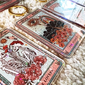 May include: A set of six clear acrylic keychains featuring tarot card designs. Each keychain has a pink border and a different tarot card image, including The Lovers, The Audacity, and The Hermit. The keychains have a glittery effect.
